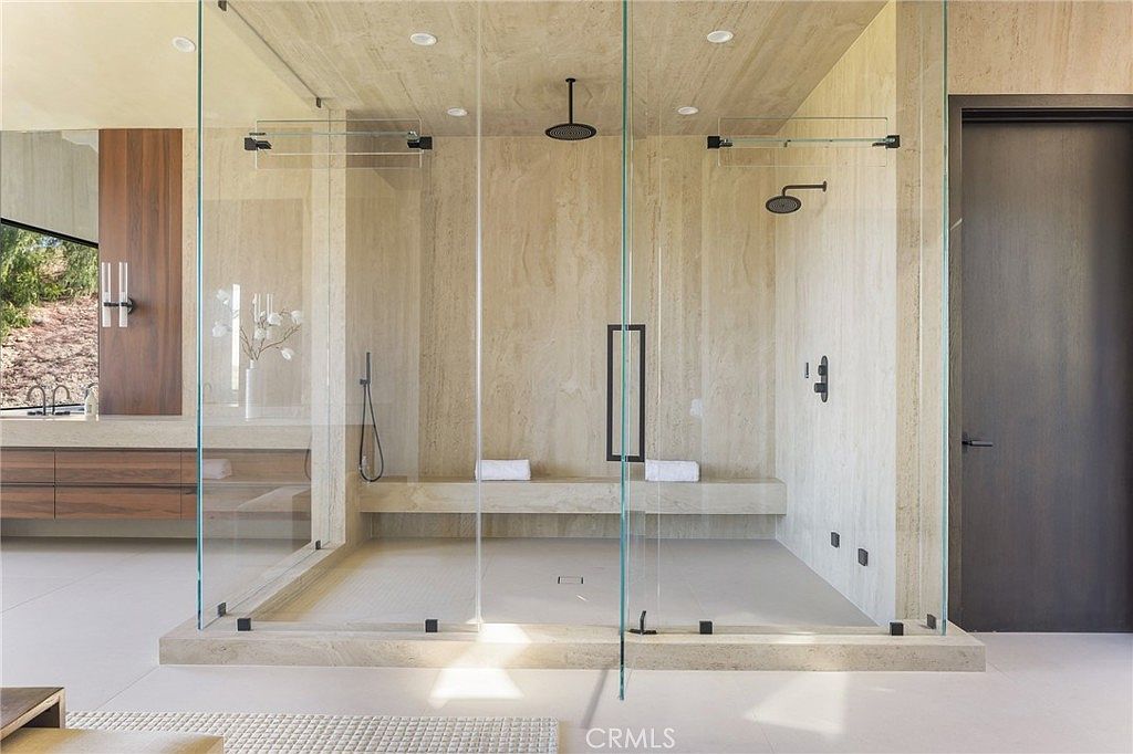 This is a luxurious primary bathroom featuring a large, walk-in shower with dual shower heads and a built-in bench. The shower is enclosed with frameless glass panels, creating a sleek and modern look. The bathroom also includes a vanity with wood cabinetry and a large mirror, enhancing the spa-like atmosphere.