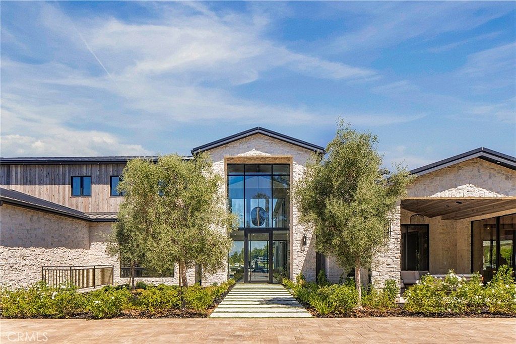 This is a front exterior view of a modern luxury home. The facade features a combination of stone and wood elements, with a striking two-story glass entryway. Landscaping includes manicured greenery and trees, enhancing the property's curb appeal and creating a welcoming atmosphere.