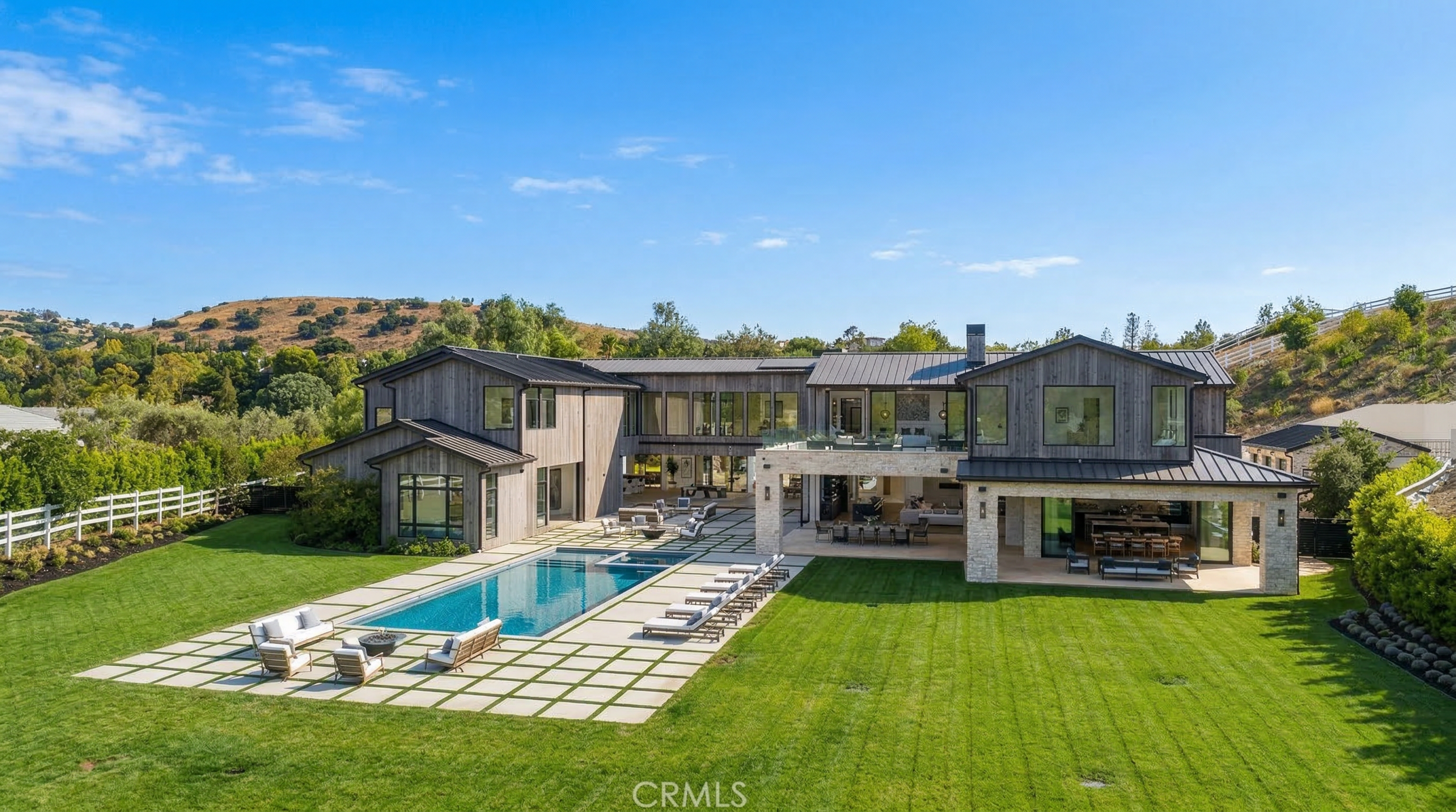 This stunning contemporary estate features a expansive rear view showcasing a custom swimming pool, a large rectangular lawn, and high-end outdoor lounging areas. The architecture boasts dark vertical wood siding contrasted by stone accents and metal roofing, creating a sophisticated farmhouse aesthetic. The integration of indoor-outdoor living spaces, including a covered patio and second-story balcony, makes this property ideal for luxury entertaining.