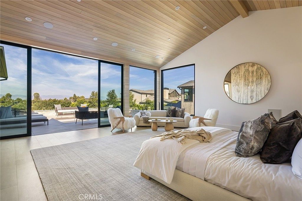 This is a bright and airy primary bedroom featuring a modern design with a vaulted wood ceiling and large sliding glass doors that open to an outdoor patio. The room includes a king-size bed with plush pillows, a seating area with stylish chairs and a sofa, and a round decorative mirror on the wall. The neutral color palette and abundant natural light create a serene and luxurious atmosphere.