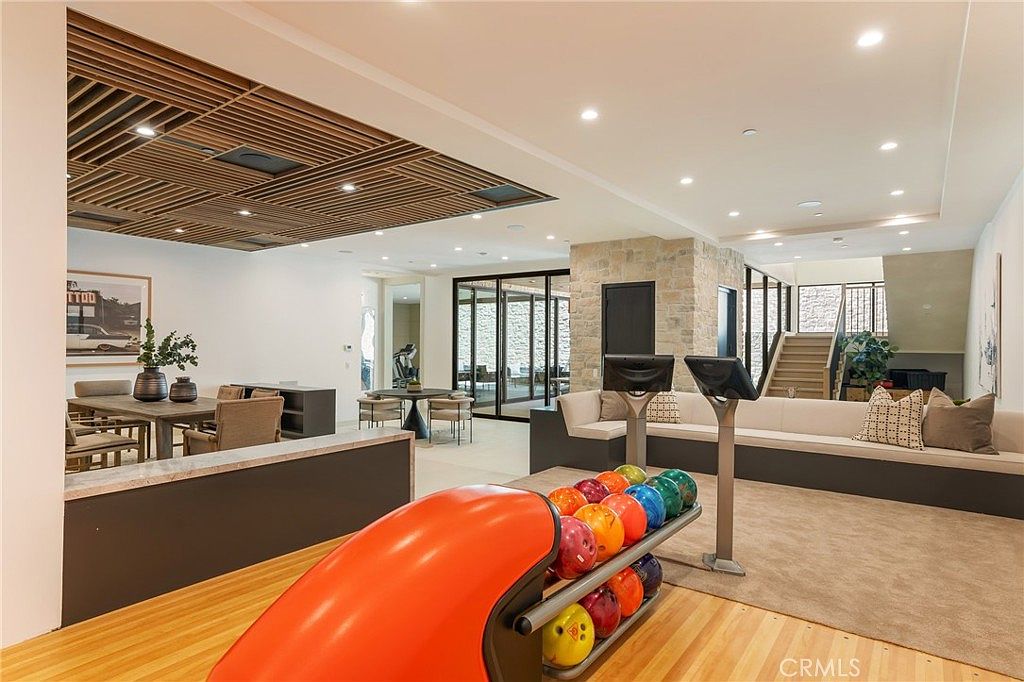 This interior shot showcases a spacious living area with a unique bowling alley feature. The room includes a dining area, a seating area with a long sofa, and a striking wooden slat ceiling design. The bowling alley adds a playful and luxurious touch to the space, making it an ideal entertainment area.