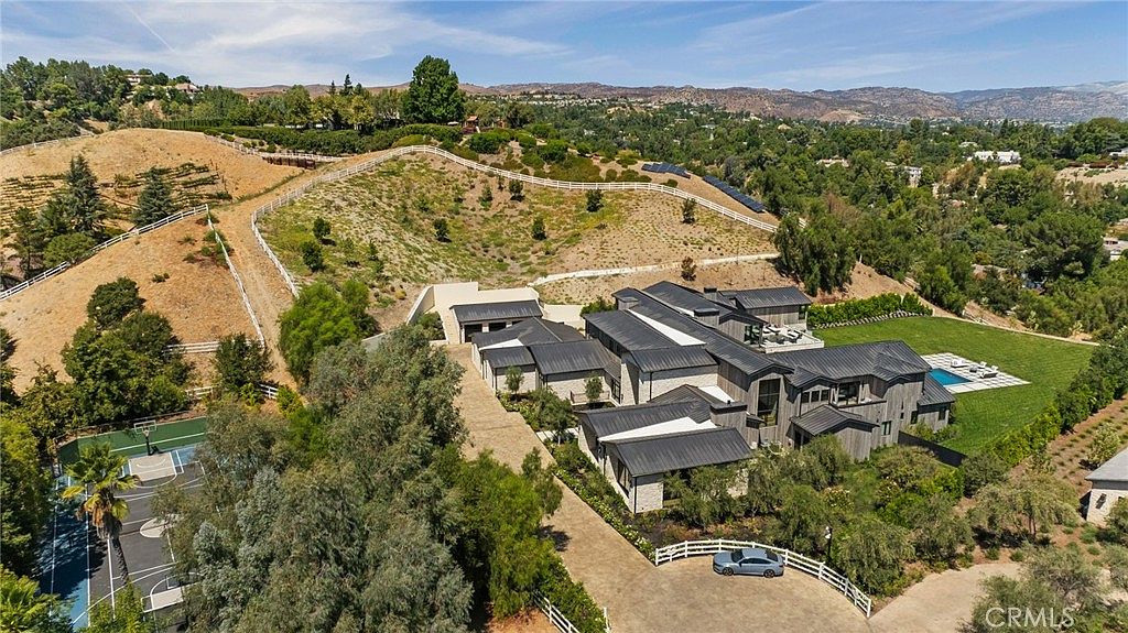 This aerial shot showcases a sprawling estate with a modern architectural style, featuring a dark roof, light stone accents, and a well-manicured lawn. A swimming pool and a basketball court are visible, along with a long driveway leading to the house. The property is surrounded by lush greenery and rolling hills, offering privacy and scenic views.