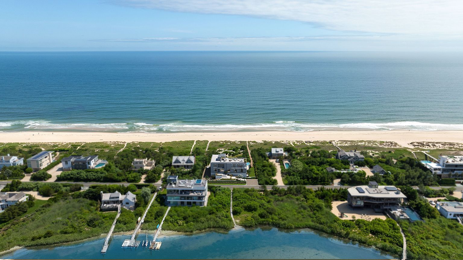 This high-angle aerial view captures a luxurious coastal enclave situated between a serene bay and the open ocean. The scene features several modern, multi-level residences nestled among lush greenery, with private docks extending into the calm water and sandy paths leading directly to the beach. The composition highlights the prime waterfront location, emphasizing the seamless integration of high-end architecture with the natural landscape.