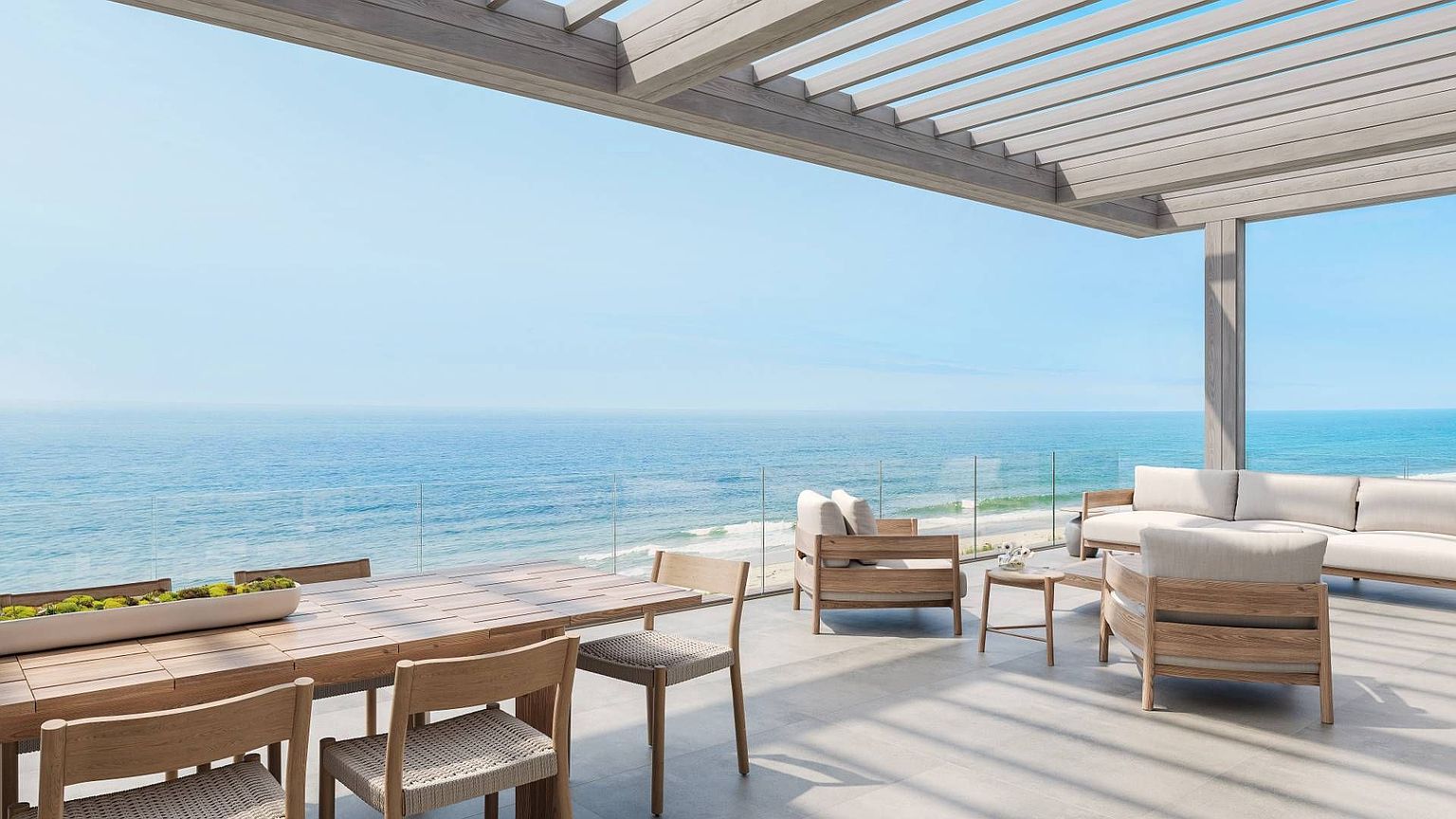 This expansive, modern outdoor terrace features a light wood pergola overhead and a seamless glass railing that provides an unobstructed, panoramic view of the ocean. The space is furnished with a light wood dining set and a comfortable lounge area with neutral-toned seating, all set against a backdrop of the horizon. The clean lines and minimalist aesthetic create a serene, high-end atmosphere perfect for coastal living.