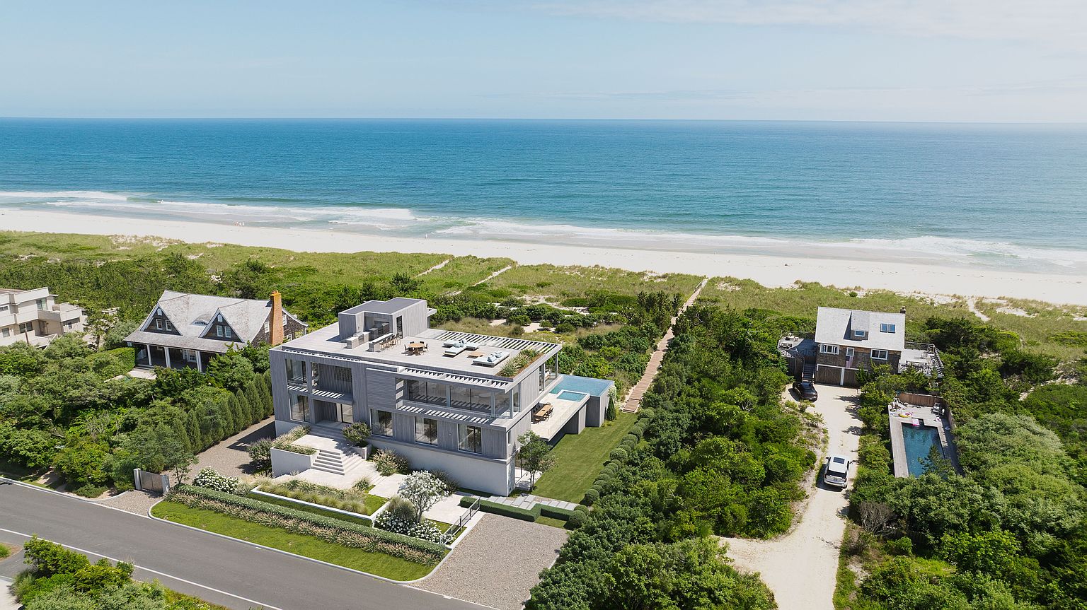This high-angle aerial perspective captures a modern, multi-level coastal residence situated directly on the beachfront. The home features a contemporary design with light-toned wood siding, a flat roof with a rooftop deck, and large windows that maximize ocean views. The property includes a private pool, manicured landscaping, and a direct wooden pathway leading from the house to the sandy beach.