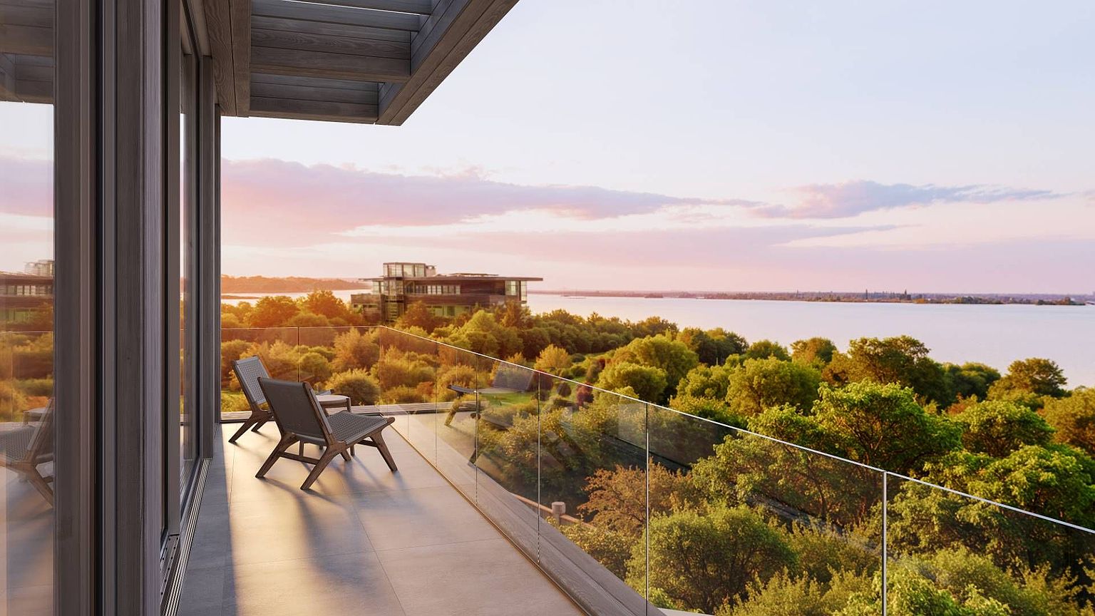 This modern balcony offers a serene, elevated perspective overlooking a lush canopy of trees and a calm body of water at sunset. The space features sleek, minimalist outdoor seating and a frameless glass railing that maximizes the expansive, scenic horizon. The warm, golden-hour light creates a tranquil and sophisticated atmosphere, highlighting the seamless transition between the indoor living area and the natural landscape.