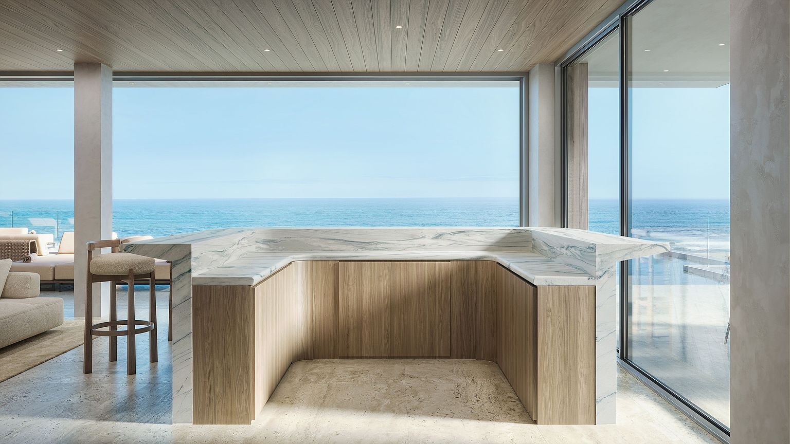 This sophisticated kitchen area features a U-shaped island with a light-toned stone countertop and warm-toned wood cabinetry. The space is framed by expansive floor-to-ceiling windows that provide an unobstructed, panoramic view of the ocean. The minimalist design, combined with the natural stone flooring and wood-paneled ceiling, creates a serene and luxurious atmosphere.
