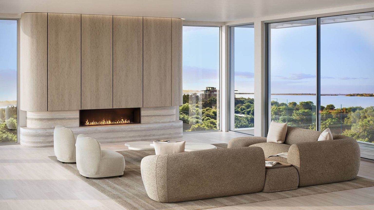 This sophisticated living room features a curved, light wood-paneled wall housing a linear fireplace set into a textured stone hearth. The space is furnished with plush, rounded modular seating and a low-profile coffee table, all arranged on a neutral area rug. Large windows provide expansive, unobstructed views of the surrounding landscape and water, creating a seamless connection between the refined interior and the natural environment.