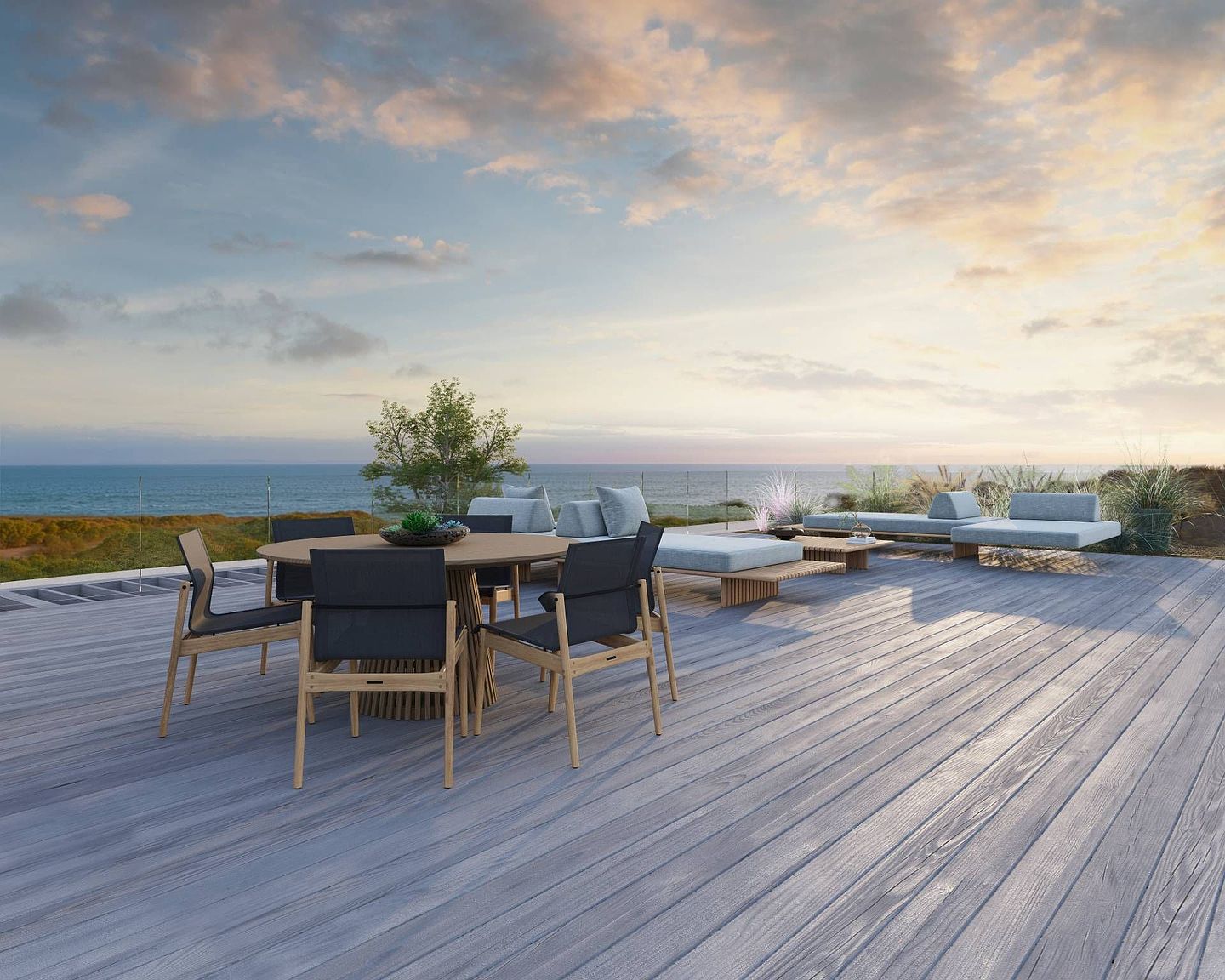 This expansive outdoor deck features a contemporary dining set with a round table and dark-upholstered chairs, complemented by low-profile lounge seating arranged for ocean views. The space is finished with light-toned wood decking and a minimalist glass railing that preserves the horizon line. The setting offers a serene, high-end atmosphere perfect for coastal living, captured from a slightly elevated perspective that emphasizes the open, airy layout.