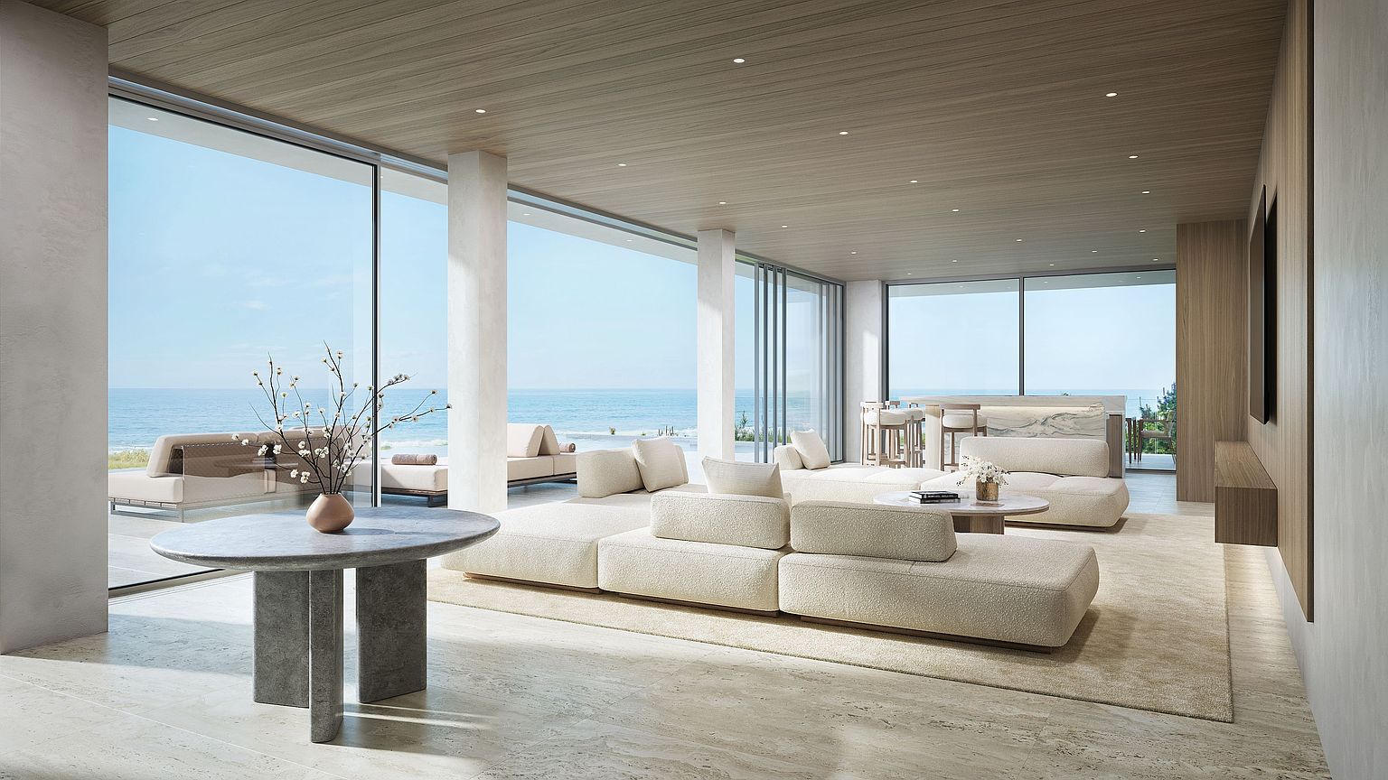 This expansive, modern living area features a minimalist aesthetic with light wood ceilings and neutral-toned stone flooring. The space is defined by floor-to-ceiling glass walls that provide panoramic ocean views and seamless access to an outdoor terrace. A large, modular cream-colored sofa serves as the centerpiece, complemented by a round stone table and a sculptural vase, creating a serene and luxurious atmosphere.