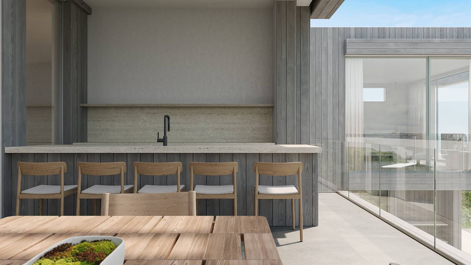 This modern outdoor balcony features a sleek bar area with a stone countertop, a minimalist faucet, and light wood cabinetry. A row of four matching light wood chairs with white cushions provides seating, while a wooden dining table with a moss-filled centerpiece sits in the foreground. The space is enclosed by glass railings, offering an open, airy feel with a view of the surrounding landscape through the large windows of the adjacent interior space.