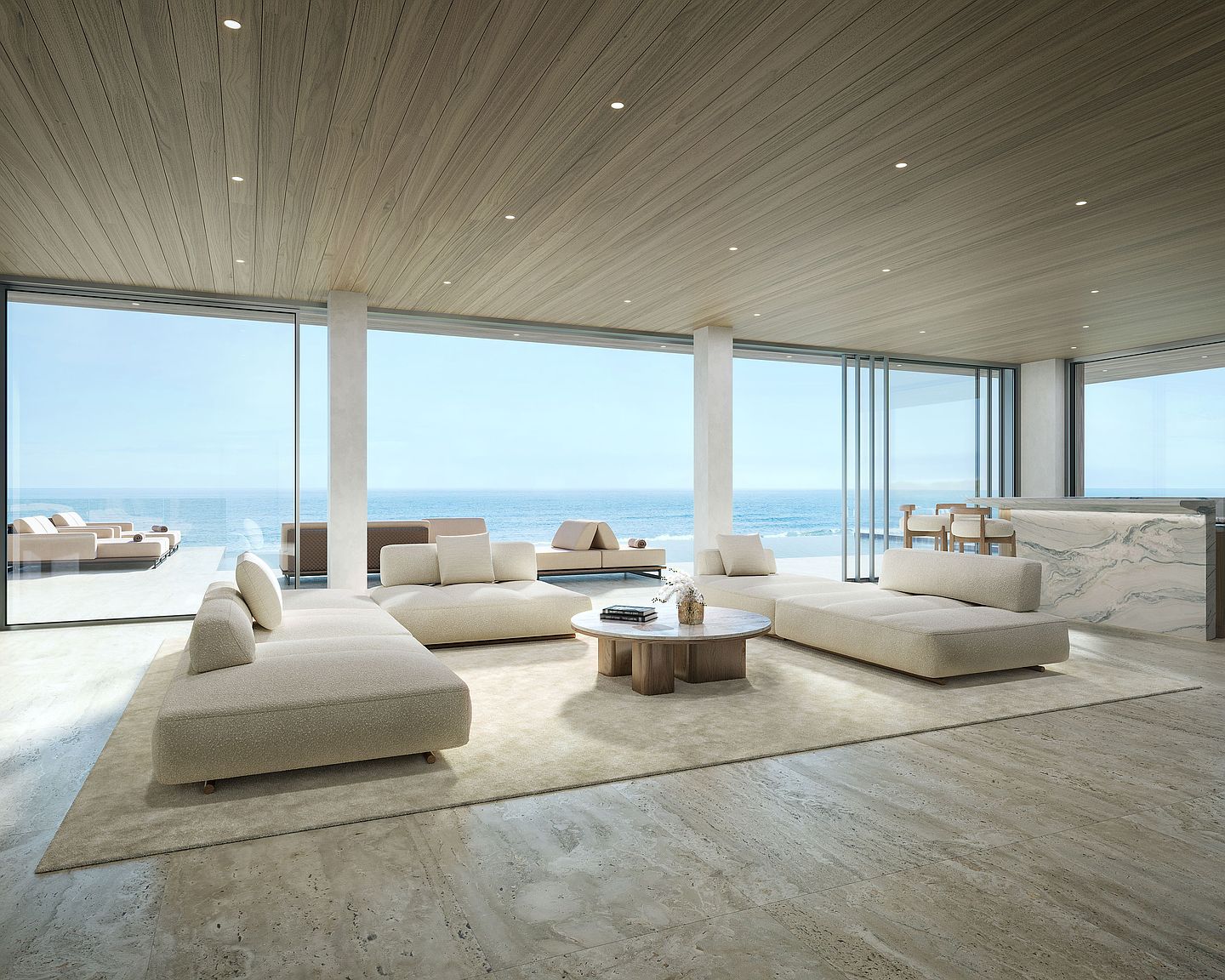 This expansive, open-concept living room features a minimalist aesthetic with light wood ceilings and large, floor-to-ceiling glass walls that frame panoramic ocean views. The space is anchored by a large, neutral-toned sectional sofa and a central stone coffee table, all resting on a soft area rug atop stone flooring. The seamless transition between the indoor living area and the outdoor terrace creates a serene, high-end coastal atmosphere.