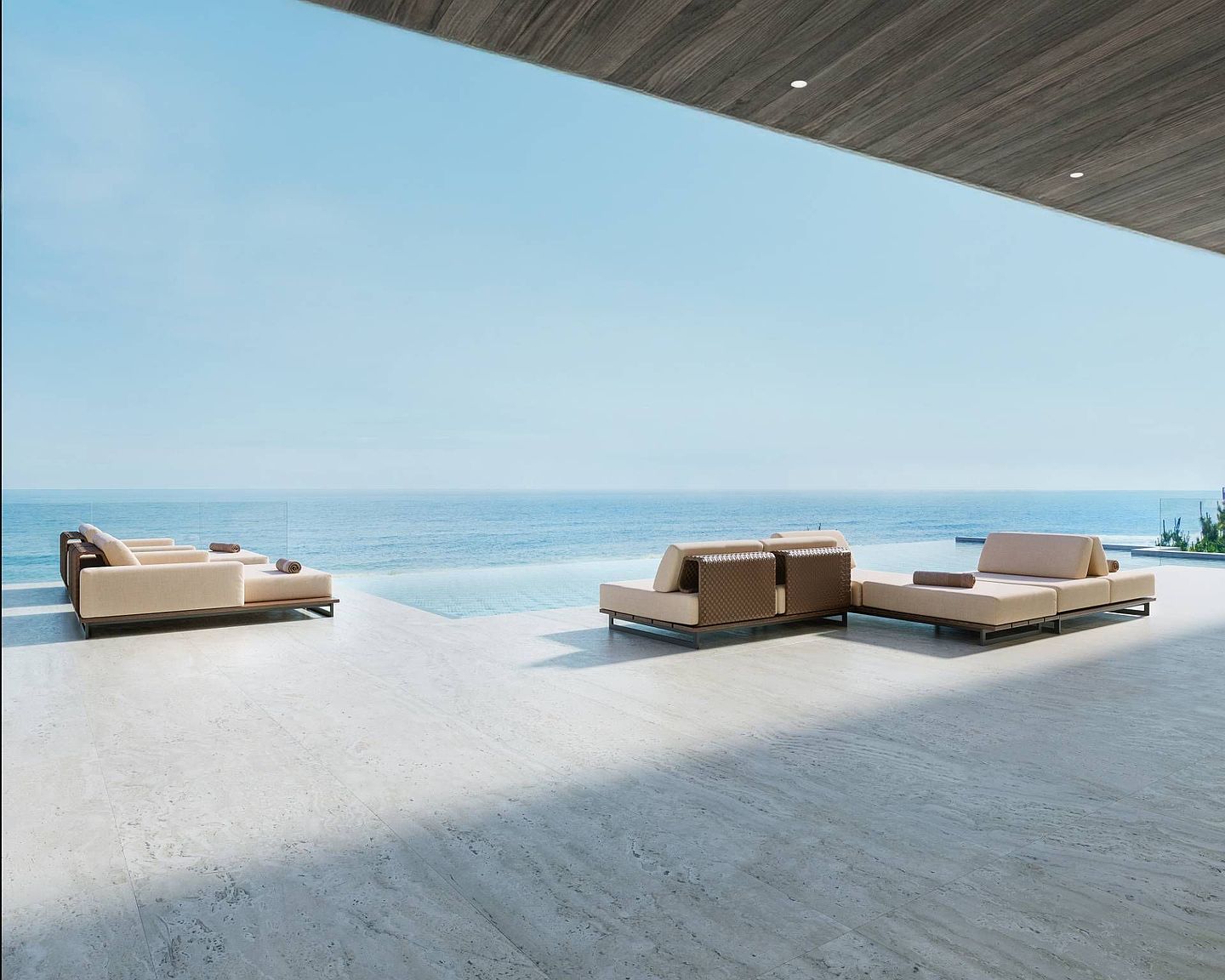 This expansive, covered outdoor terrace features sleek, light-toned stone flooring and a minimalist design that seamlessly transitions into an infinity-edge pool. The space is furnished with modern, low-profile lounge seating arranged to maximize the panoramic ocean views. The wide, open-air perspective emphasizes a luxurious, serene atmosphere perfect for high-end coastal living.
