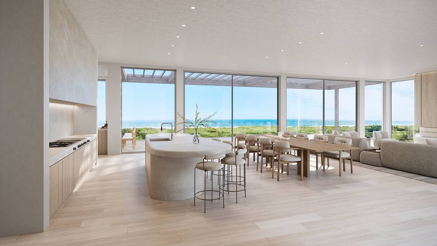 This open-concept living space features a minimalist kitchen with a curved stone island and light wood cabinetry. Large glass panels provide an expansive, unobstructed view of the ocean and coastal landscape, bathing the room in natural light. The dining area and living room flow seamlessly from the kitchen, creating a bright, airy atmosphere with a modern, coastal aesthetic.