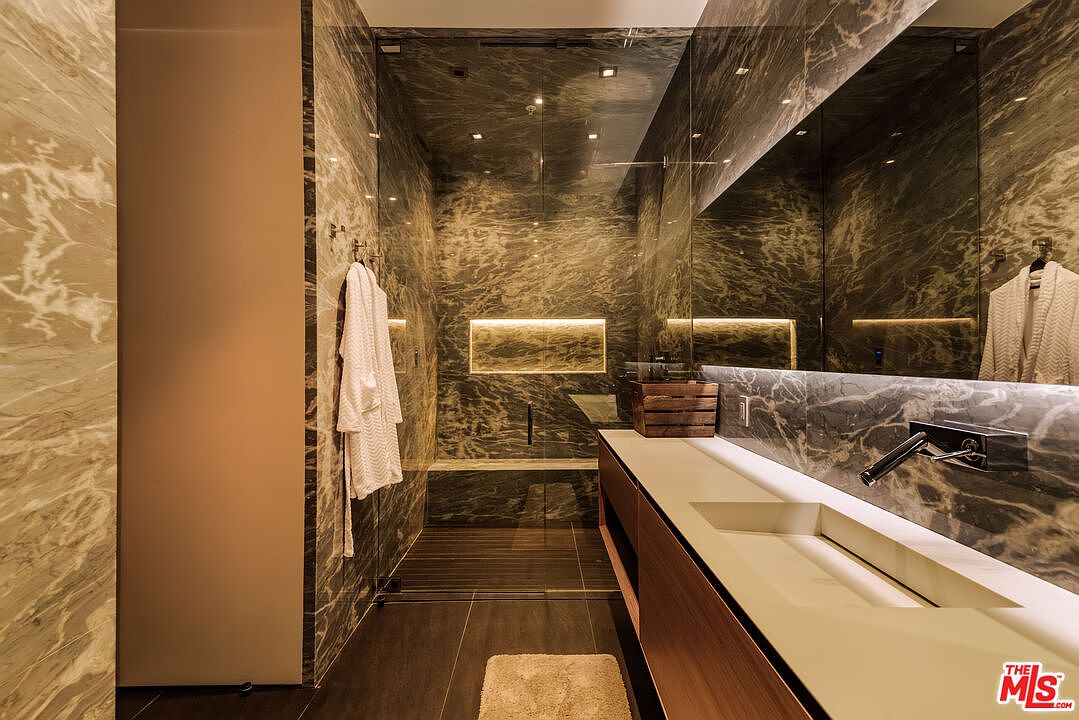 This luxurious, modern bathroom features dramatic, dark-veined marble walls and a sleek, minimalist vanity with a wall-mounted faucet. A glass-enclosed walk-in shower with integrated lighting and a built-in bench creates a spa-like atmosphere, while a large mirror enhances the sense of depth. The overall aesthetic is sophisticated, high-end, and contemporary.