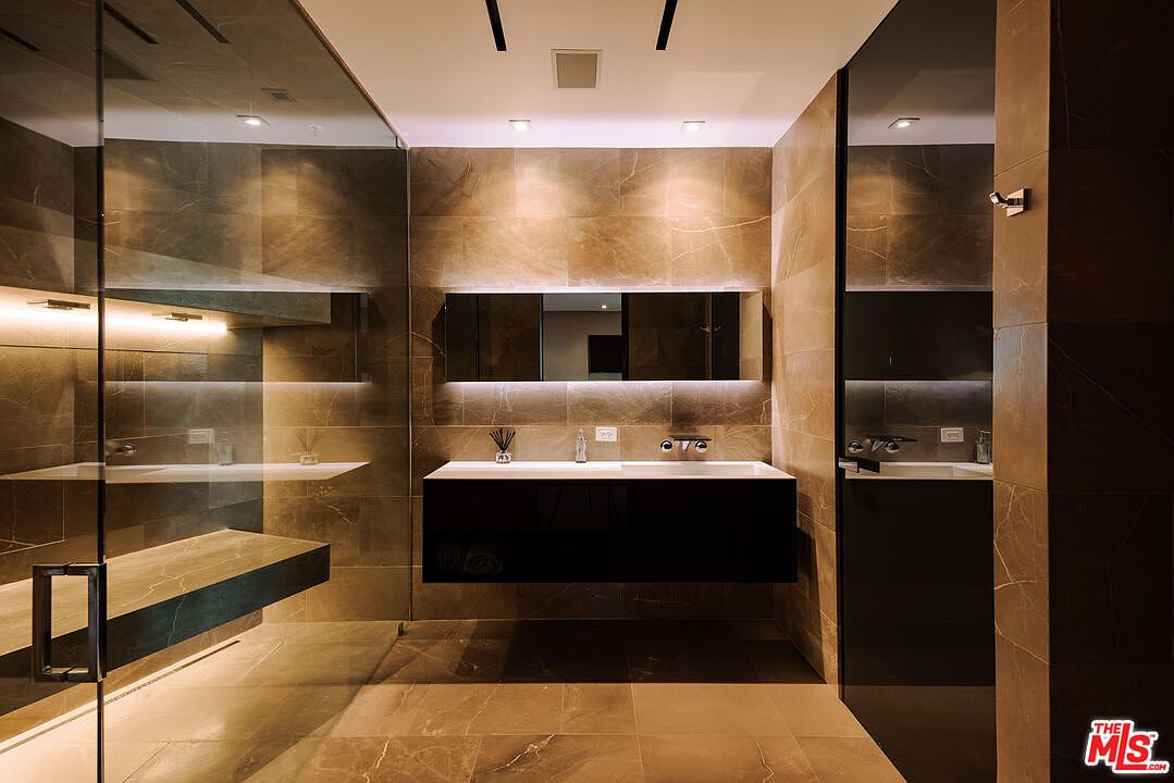 This luxurious, modern primary bathroom features floor-to-ceiling brown marble tiling and a sleek, floating black vanity with a minimalist sink. A large, frameless glass-enclosed shower area sits to the left, while a wide, backlit mirror creates a sophisticated, spa-like atmosphere. The perspective is centered, emphasizing the clean lines and high-end finishes of this contemporary design.
