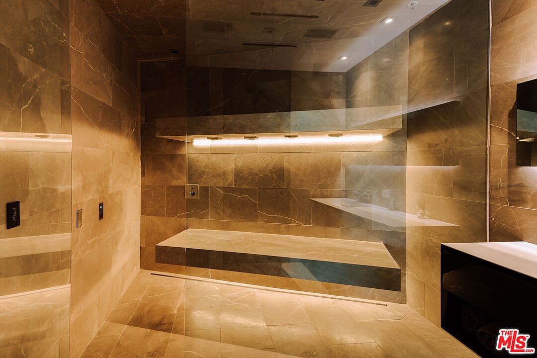 This luxurious, spa-inspired walk-in shower features floor-to-ceiling marble tiling and a sleek, floating stone bench. The space is illuminated by warm, integrated LED strip lighting beneath a floating shelf, creating a sophisticated and tranquil atmosphere. A large glass partition provides a clean, modern aesthetic, highlighting the high-end finishes and minimalist design.
