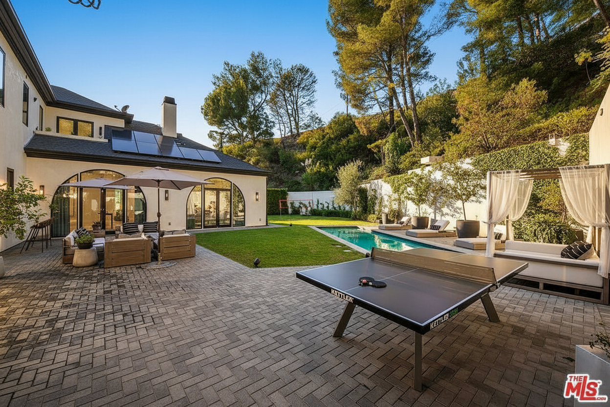 This image showcases the rear exterior of a luxurious home with a well-manicured backyard. A patio features outdoor seating, a ping pong table, and a poolside cabana, suggesting an entertainment-friendly space. The landscape includes a swimming pool, grassy area, and lush hillside, contributing to a resort-like atmosphere.