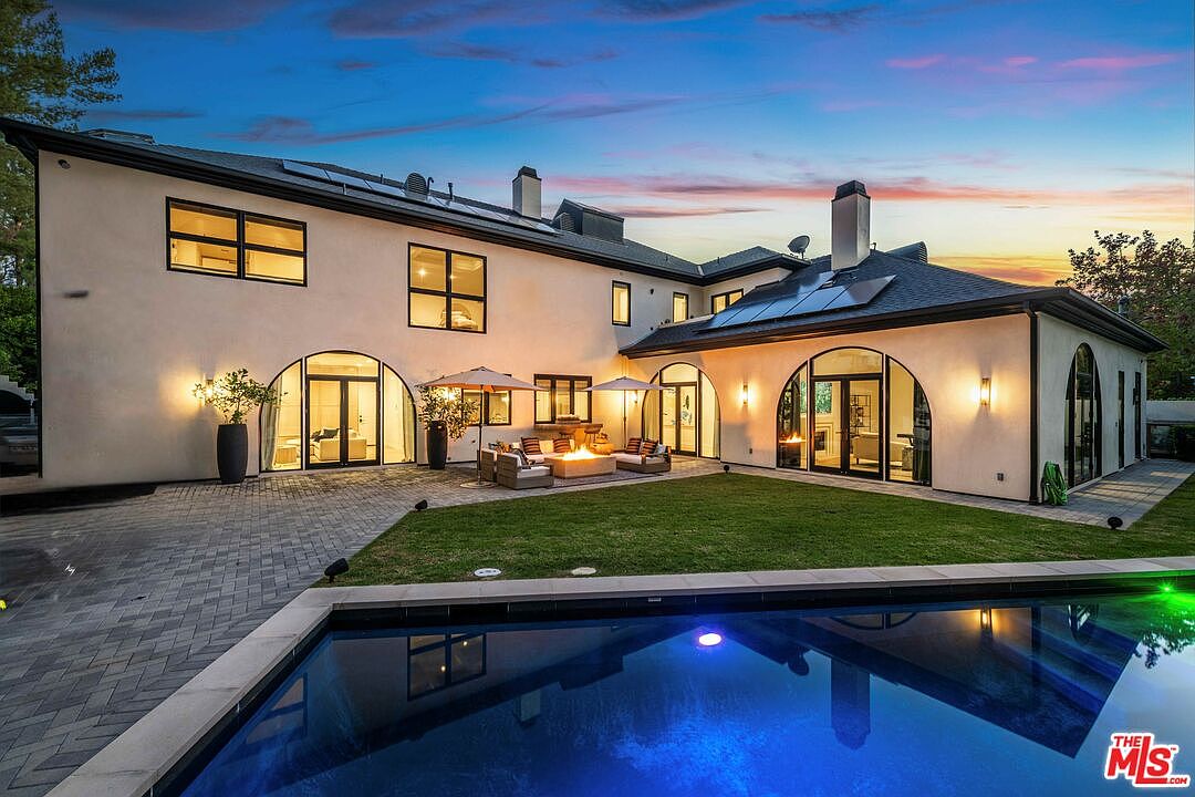 This is a rear exterior view of a luxurious home featuring a modern design with arched windows and doors. A rectangular pool with blue water is in the foreground, reflecting the house and the colorful sky at dusk. The backyard includes a well-maintained lawn and a patio area with a fire pit and outdoor seating, creating an inviting space for relaxation and entertainment.