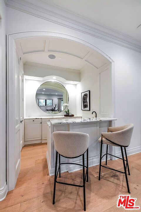 This interior shot showcases a stylish kitchen bar area with two modern bar stools. The bar features a marble countertop and white cabinetry, complemented by a large round mirror and an elegant archway. The space is well-lit and exudes a sophisticated, clean aesthetic.