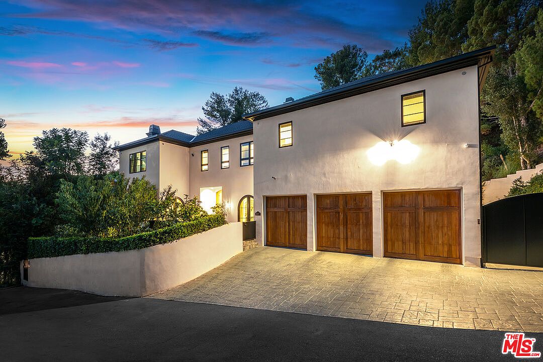 This is a front exterior view of a two-story house at dusk. The house features a light-colored stucco exterior, dark trim, and three wooden garage doors. The driveway is paved with interlocking stones, and the landscaping includes a manicured hedge and mature trees. The sky is a mix of blue, pink, and orange, creating a warm and inviting atmosphere.