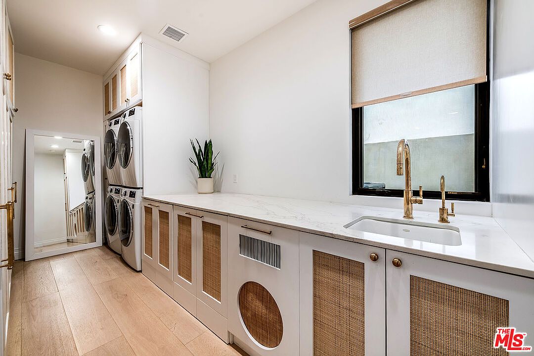 This is a bright and modern laundry room featuring stacked washer and dryer units, custom cabinetry with woven panel inserts, and a marble countertop with a sink and gold fixtures. A large window provides natural light, and the light wood flooring adds warmth to the space. The room appears clean and well-organized, suggesting a functional and stylish design.