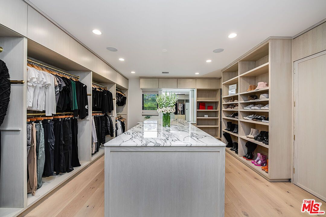 This is a spacious walk-in closet featuring custom shelving and hanging rods for clothing and shoes. A central island with a marble countertop provides additional storage and display space. The closet is well-lit with recessed lighting and has a clean, organized aesthetic, enhancing the appeal for potential buyers.