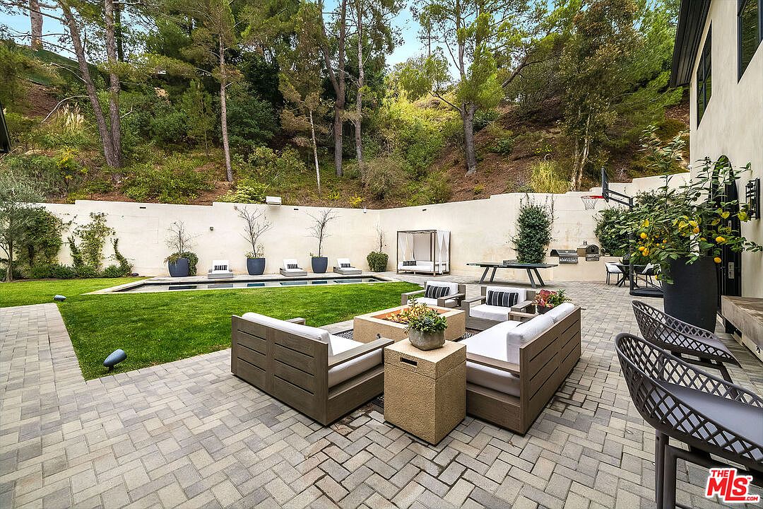 This image showcases a beautifully landscaped backyard featuring a pool, a well-maintained lawn, and a stylish patio area. The patio is furnished with modern outdoor seating arranged around a fire pit, creating an inviting space for relaxation and entertainment. The surrounding greenery and tall trees provide privacy and a serene atmosphere.