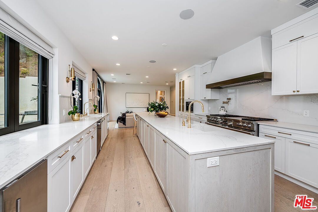 This is a bright, modern kitchen with white cabinetry, marble countertops, and stainless steel appliances. A large island dominates the center of the room, featuring a gold faucet and a bowl of fruit. The kitchen seamlessly transitions into a living area, creating an open and inviting space.