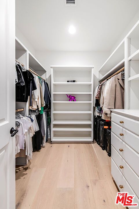 This is a well-organized walk-in closet featuring custom shelving and drawers. The closet is brightly lit and contains a variety of clothing items neatly arranged on hangers and shelves. A central shelving unit provides additional storage space, and the light wood flooring adds warmth to the space.
