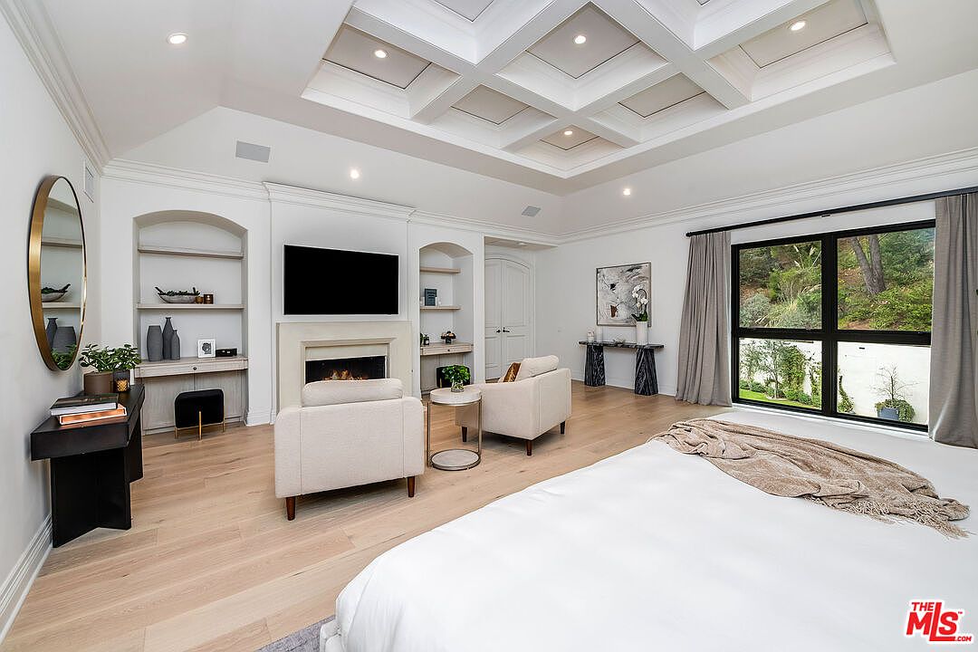 This is a bright and spacious primary bedroom featuring a coffered ceiling, a fireplace with a mounted TV, and built-in shelving units. The room is furnished with two armchairs, a side table, and a large bed with a throw blanket. A large window provides natural light and a view of the outdoors, enhancing the room's serene and luxurious atmosphere.