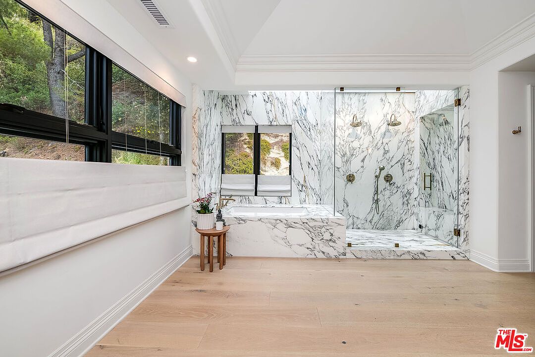 This is a luxurious primary bathroom featuring marble walls and flooring, a freestanding tub, and a glass-enclosed shower with dual shower heads. Large windows provide natural light and views of the surrounding greenery. The overall impression is one of elegance and spa-like tranquility.