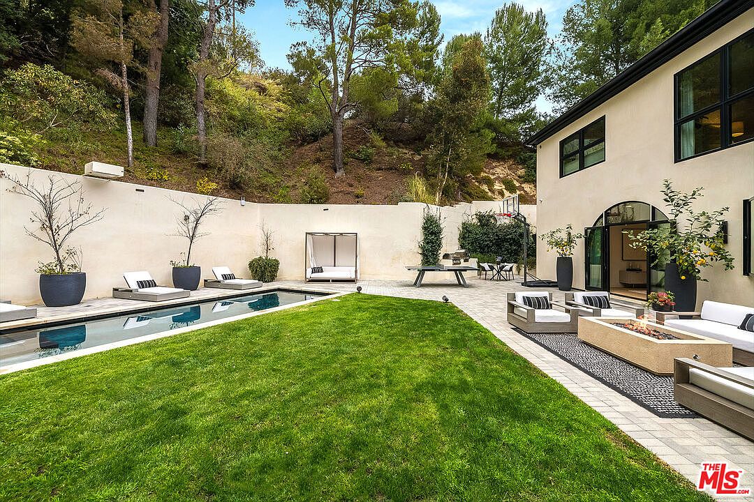This image showcases a beautifully landscaped backyard featuring a modern pool, lush green lawn, and a stylish patio area. The patio includes comfortable seating around a fire pit, creating an inviting outdoor living space. The high wall provides privacy and a sense of seclusion, while the mature trees in the background add to the natural ambiance.