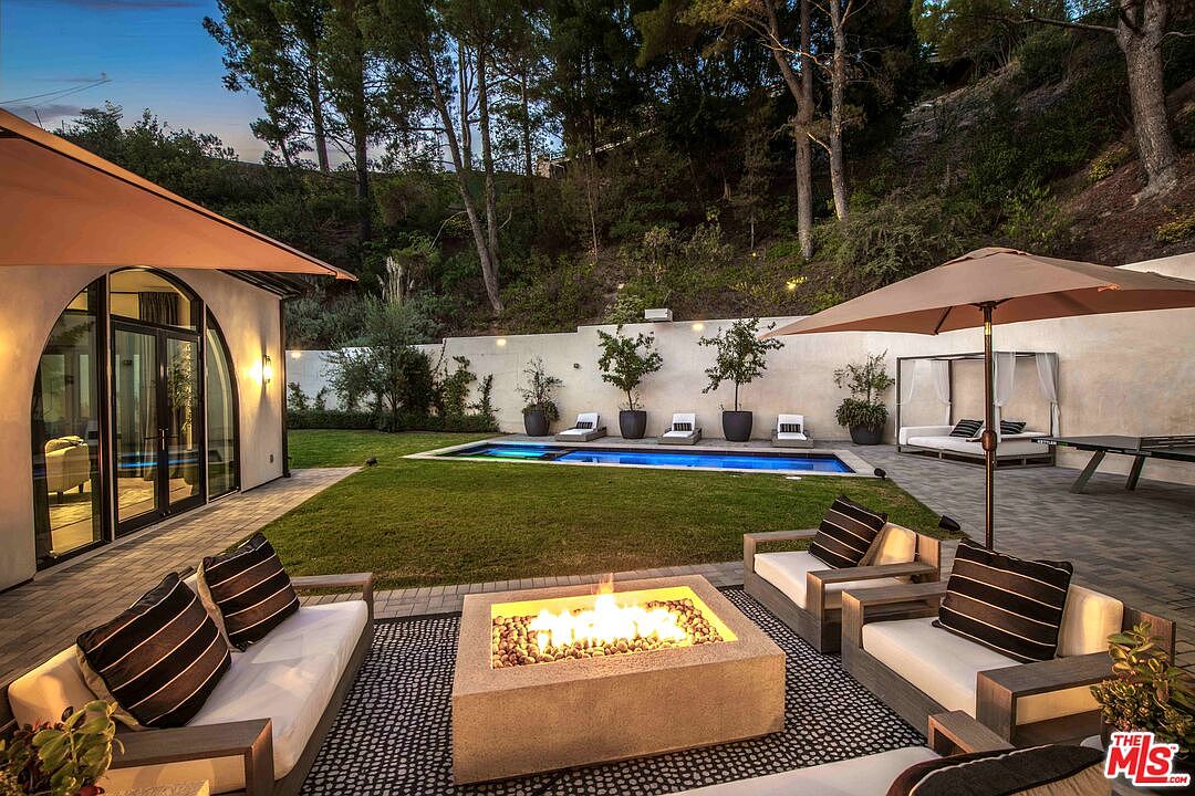 This is a beautifully landscaped backyard featuring a modern fire pit lounge area, a swimming pool, and a covered daybed. The yard is well-maintained with lush green grass and mature trees providing privacy. The overall impression is one of luxury and relaxation, perfect for outdoor entertaining.