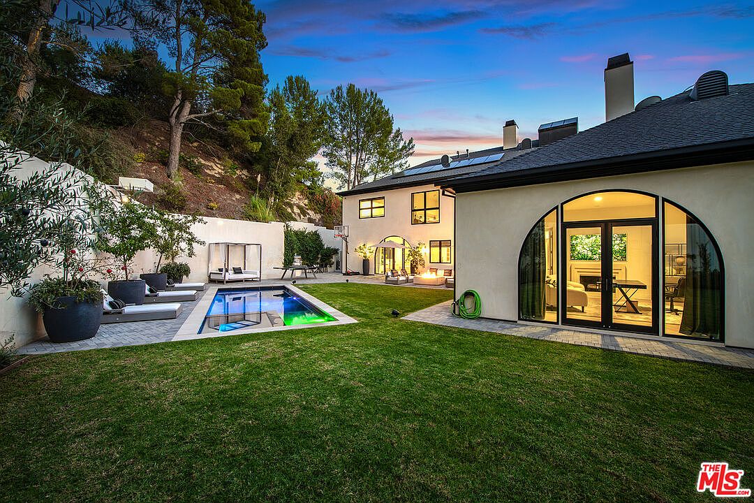 This image showcases a beautifully landscaped backyard featuring a modern pool with vibrant lighting, surrounded by lush green grass and manicured plants. The property includes a stylish outdoor lounge area and a contemporary home with a unique arched window, creating an inviting and luxurious outdoor living space. The overall impression is one of tranquility and upscale design.
