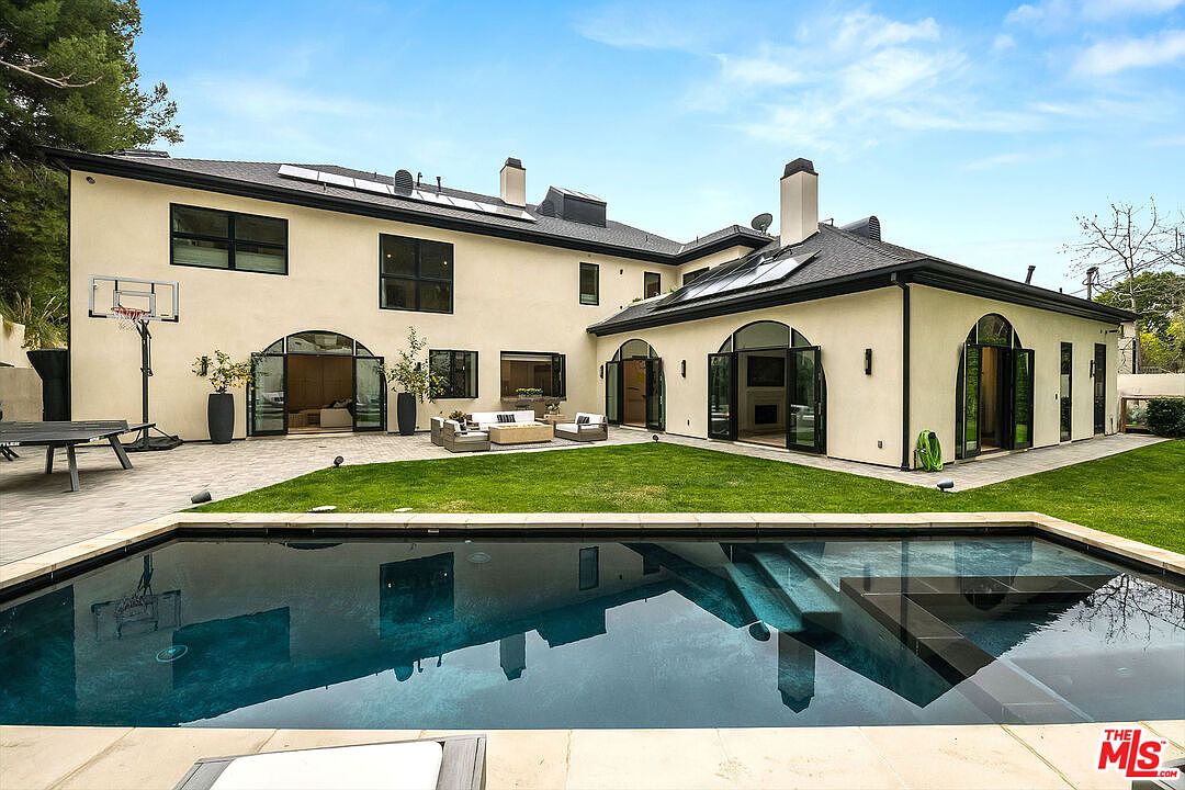This image showcases the rear exterior of a luxurious home, focusing on the pool and backyard area. The pool features clear blue water and integrated steps, reflecting the architecture of the house. The backyard includes a well-maintained lawn, a patio area with outdoor seating, and the home's elegant facade with arched doorways and dark-framed windows, creating an inviting and upscale outdoor living space.