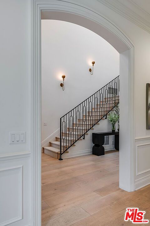 This interior shot showcases a bright hallway with a staircase. The staircase features a black metal railing and carpeted steps, illuminated by sconces on the wall. The hallway has light wood flooring and white walls, creating a clean and elegant aesthetic.
