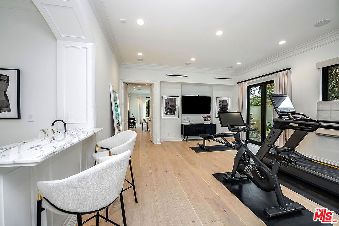 This is an interior shot of a home gym featuring modern exercise equipment, including a treadmill and stationary bike. The room has light hardwood floors, white walls, and recessed lighting, creating a bright and clean workout space. A bar area with seating is visible on the left, adding a unique touch to the gym.