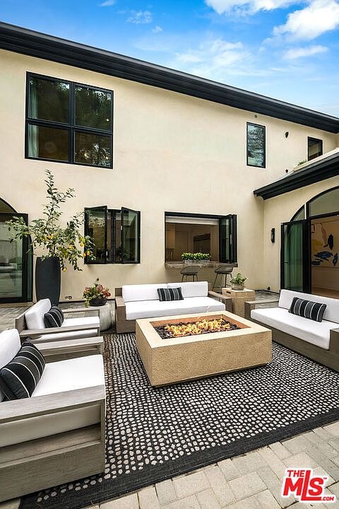 This is a view of an outdoor patio area featuring modern furniture, including sofas and a fire pit. The patio is decorated with a black and white patterned rug, and the surrounding architecture is contemporary with large windows and doors. The space appears inviting and designed for relaxation and entertainment.