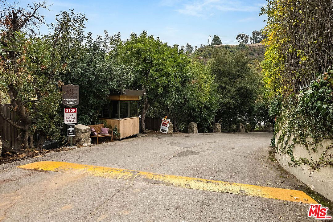 The image showcases the entryway to a property, featuring a guardhouse with glass windows and wooden siding. A 'STOP FOR VEHICLE CHECK' sign is visible, along with a bench and lush greenery surrounding the area. The asphalt driveway leads into the property, creating a secure and private entrance.