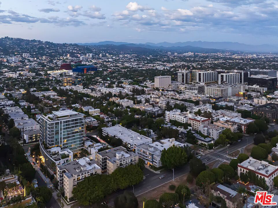 This aerial view captures a densely populated urban environment, likely a residential area in a city. Buildings of various sizes and architectural styles are visible, interspersed with mature trees and streets with light traffic. The image provides a sense of the overall layout and urban landscape of the real estate location.