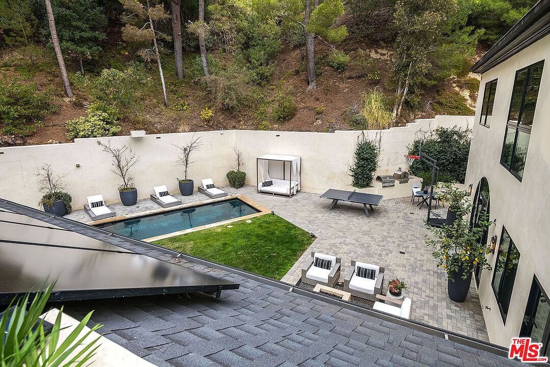 This image showcases a well-designed backyard featuring a rectangular pool, a small patch of grass, and a paved patio area. The yard is enclosed by a high white wall, providing privacy, and is surrounded by lush greenery. The patio includes lounge chairs, a daybed, a ping pong table, and a seating area, creating an inviting outdoor living space.