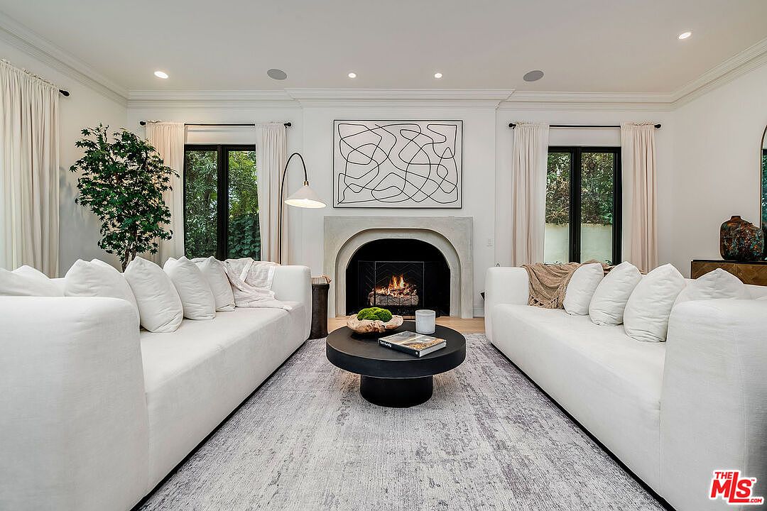 This is a bright and airy living room featuring two white sofas facing each other, separated by a round black coffee table. A modern fireplace with abstract art above it serves as a focal point, flanked by windows with light-colored curtains. The room is well-lit with recessed lighting and natural light, creating a welcoming and sophisticated atmosphere.