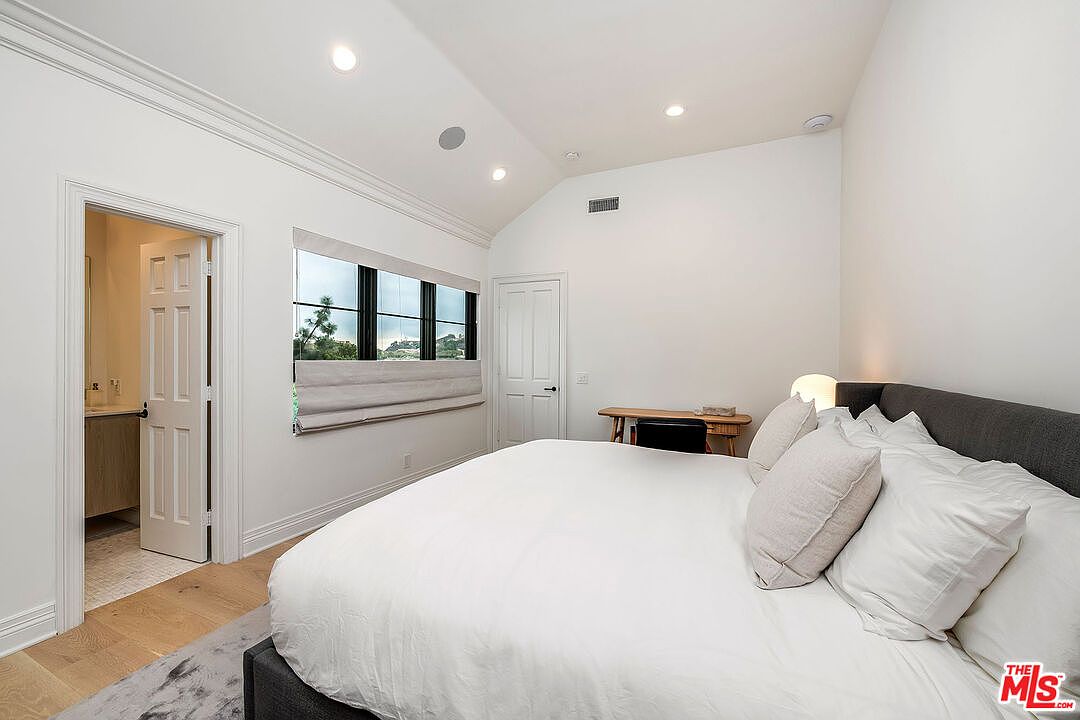 This is a bright and airy bedroom featuring a large bed with white linens and neutral-toned pillows. The room has a minimalist aesthetic with white walls, a small desk, and a window with a view. The hardwood floors and recessed lighting add to the modern feel of the space.