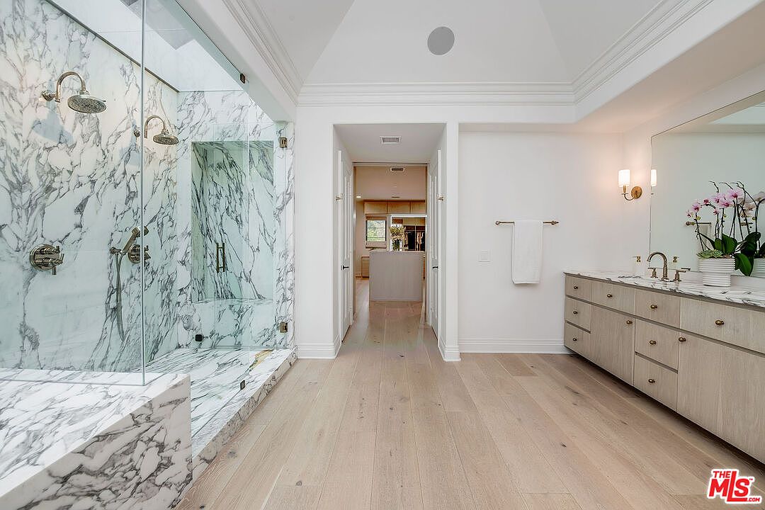 This is a luxurious primary bathroom featuring a marble-clad walk-in shower with dual shower heads and glass enclosure. The bathroom boasts light wood flooring, a double vanity with marble countertop, and ample storage. The overall aesthetic is bright, spacious, and modern, creating a spa-like atmosphere.