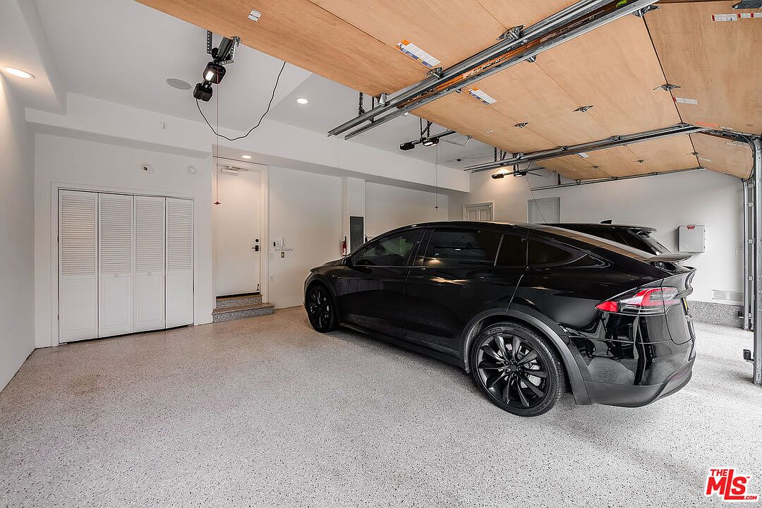 This is an interior shot of a well-lit garage featuring a sleek, black SUV parked inside. The garage has a clean, modern aesthetic with white walls, a speckled floor, and a unique wooden garage door design. A set of white bi-fold doors and an entry door are visible on the left, adding to the functionality of the space.