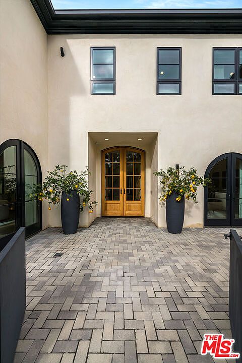 This image showcases an elegant entryway with a herringbone brick pathway leading to double wooden doors. Flanking the doors are arched windows and large potted lemon trees, adding a touch of sophistication and curb appeal. The exterior is finished in a neutral tone, complemented by black window frames and trim.
