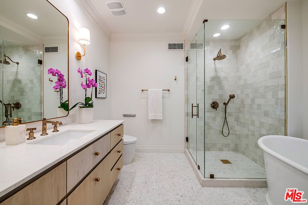 This is a well-lit primary bathroom featuring a modern design with a double vanity, a large mirror, and a glass-enclosed shower. The vanity has light wood cabinets and a white countertop, complemented by bronze fixtures. The shower showcases light-colored tile and bronze hardware, while a freestanding bathtub adds a touch of luxury.
