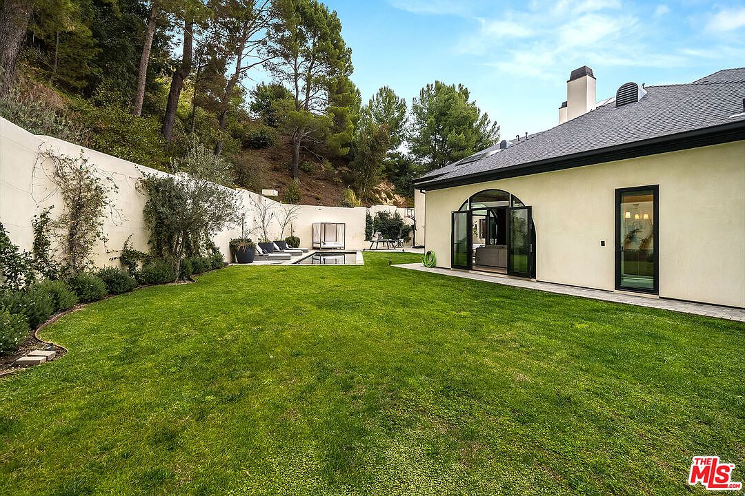 This image showcases a well-manicured backyard featuring a lush green lawn, a modern swimming pool with lounge chairs, and a stylish cabana. The property is enclosed by a high wall, providing privacy, and the house itself has large glass doors that open onto the yard, creating a seamless indoor-outdoor living experience. The overall impression is one of luxury and tranquility.