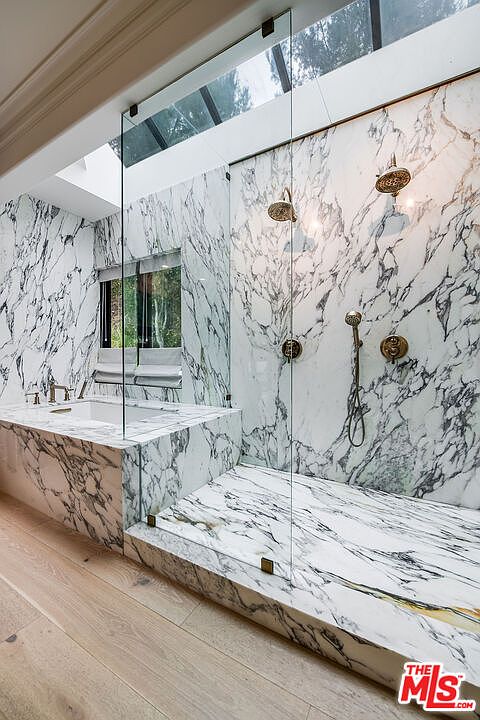 This is a luxurious primary bathroom featuring marble walls and flooring, a glass-enclosed shower with dual shower heads, and a soaking tub. The natural light from the skylight enhances the elegant and spa-like atmosphere. The perspective is from the entrance of the bathroom, showcasing the entire space.